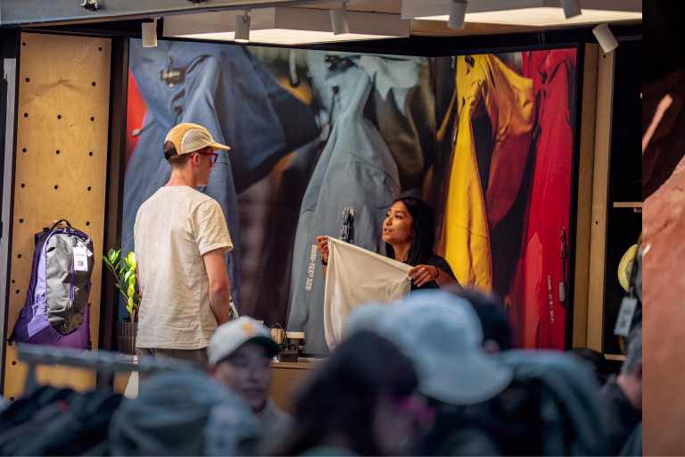 A store staff member holds up a white garment for a customer inside an outdoor apparel pop-up, with a large backdrop of colorful rain jackets and backpacks on display around them.