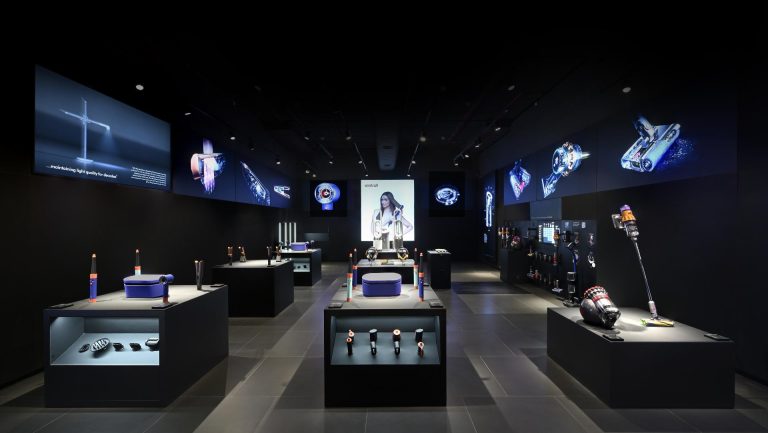 A sleek, darkened Dyson showroom with spotlit product pedestals displaying hair tools, vacuums, and accessories, surrounded by glowing blue product screens on the walls and ceiling.