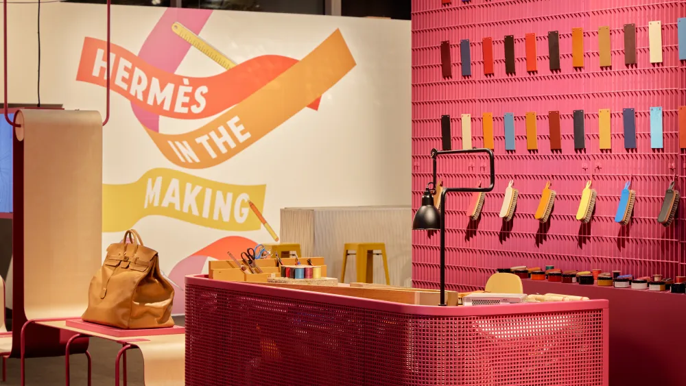 Interior of the Hermès "In the Making" pop-up workshop with a raspberry pink workstation, a wall of colorful leather swatches, craft tools, and a bold illustrated mural on the wall.