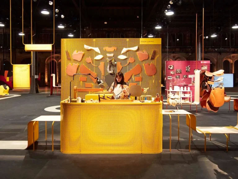 An artisan works at a bold yellow mesh workstation decorated with scattered leather cut-outs in a large exhibition hall, with a raspberry pink craftsmanship station and suspended saddle pieces visible in the background.